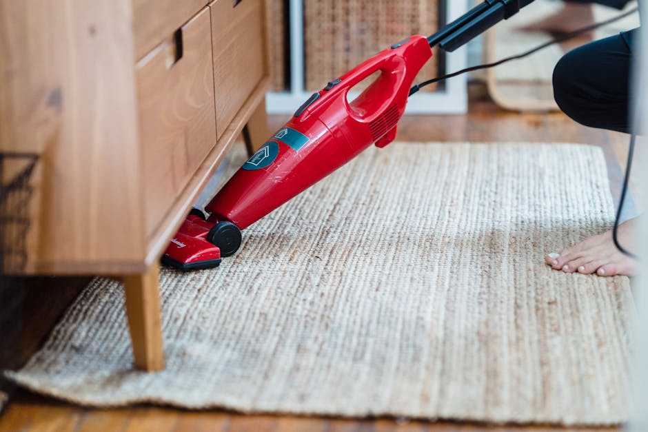 A person using a red handheld vacuum cleaner on a beige textured area rug in a living room, with visible wooden furniture on the left and a patterned wooden floor beneath. The vacuum is positioned nea