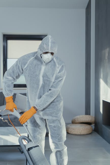 A professional cleaner wearing a white protective coverall, safety mask, and orange gloves is operating a vacuum or cleaning device on a light-colored tiled floor in a modern, well-lit room with large